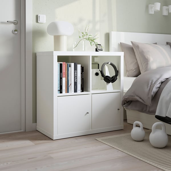 White KALLAX cube storage unit with books, headphones, and kettlebells; small, minimalist bedroom with light-colored flooring.