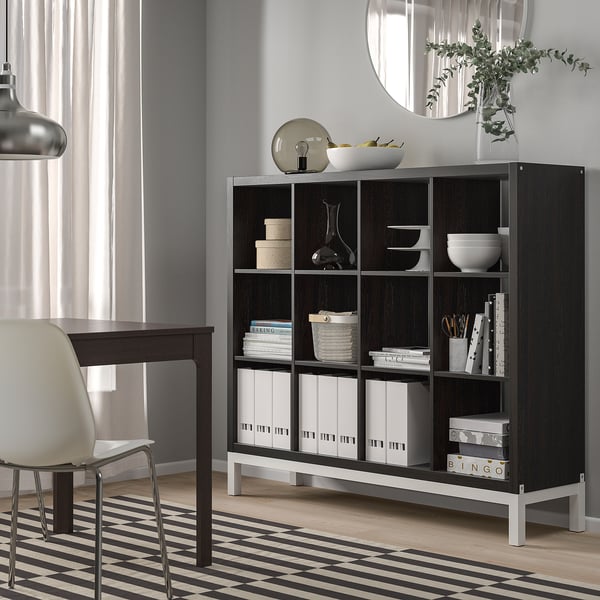 The KALLAX black shelving unit holds various items, standing tall next to a desk and chair, with neatly organised books and decor.