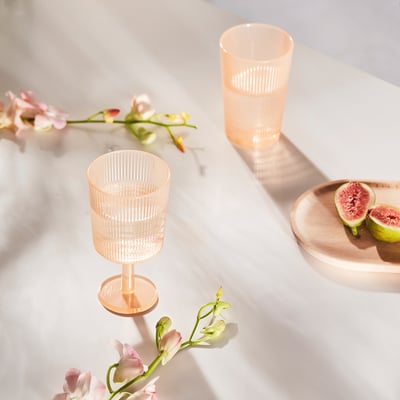 Two pink plastic wine glasses, one strawberry on a wooden plate, surrounded by flowers on a marble table.