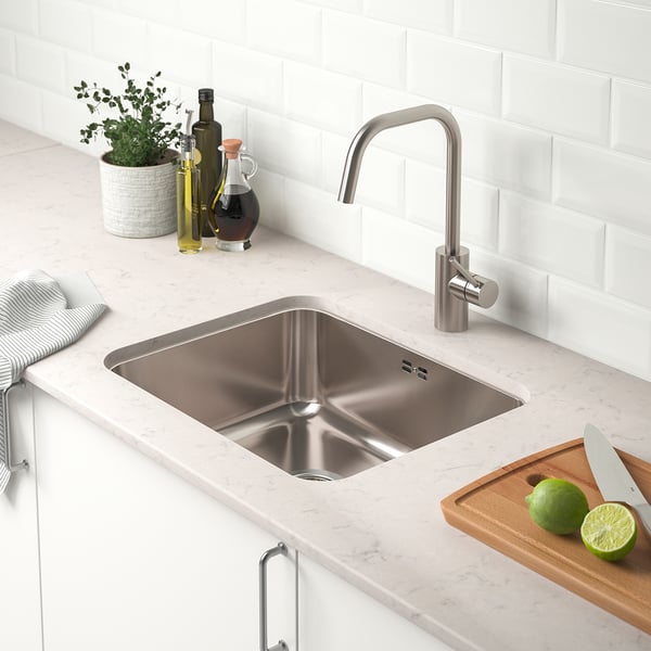 Kitchen sink with tap, knives, limes, countertop with bottles, plant, and cutting board nearby.