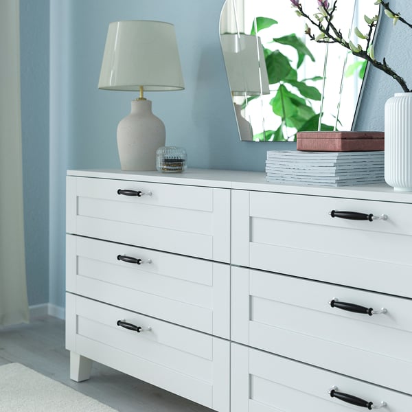 White dresser with dark accents, six drawers, lamp, decor, and houseplant against blue wall.
