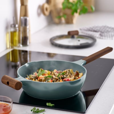 KLIPPFISK pan with stir-fry on stove, wooden handles, glass lid, cutting board & bottles in background.