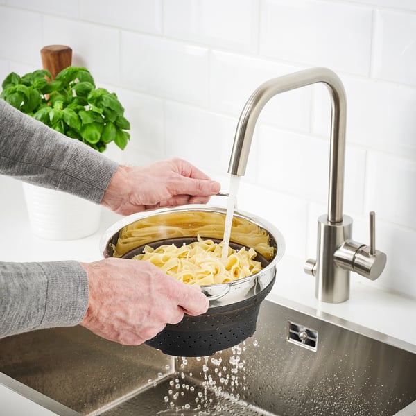 Person rinses pasta in stainless steel sink using black collapsible colander under modern silver tap.