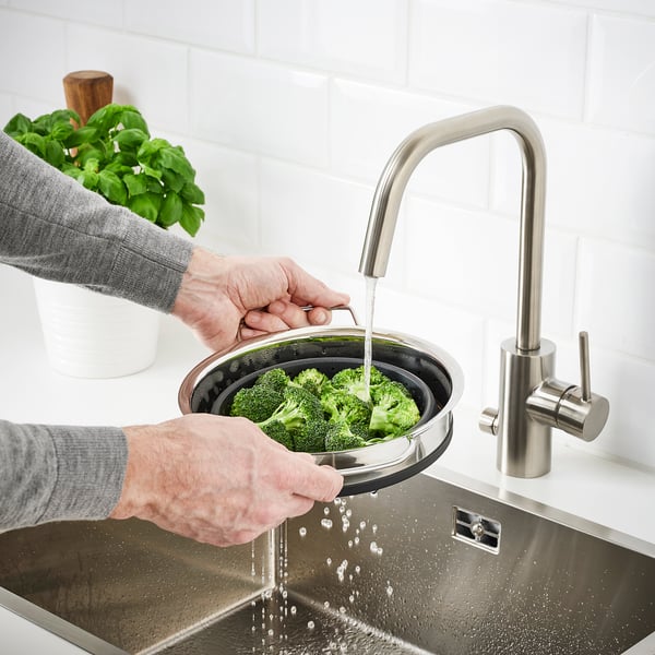 Person washes broccoli in a sink using a foldable colander attached to a modern tap.