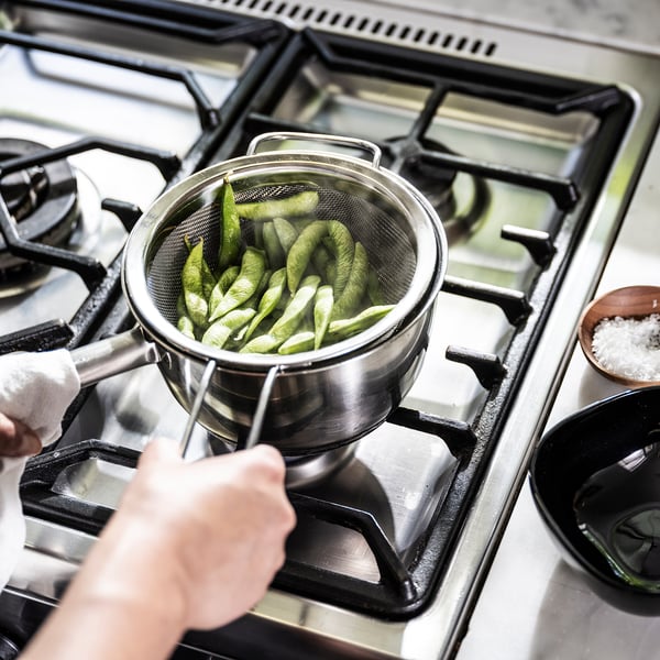 A hand holds a silver KLOCKREN colander with green fruits over a gas stove.
