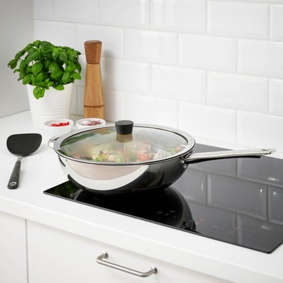 Stainless frying pan with glass lid simmers food on black cooktop near white brick wall.