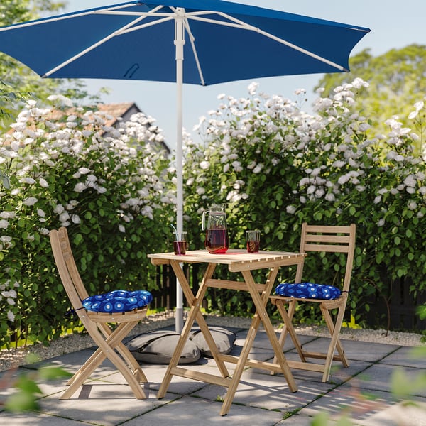 In a lush garden, a small wooden table with a blue umbrella holds drinks. Two wooden chairs with blue cushions face blooming white flowers.