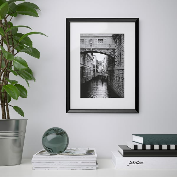 Black framed venice canal on wall; below it, stack of magazines with green glass sphere and plant.