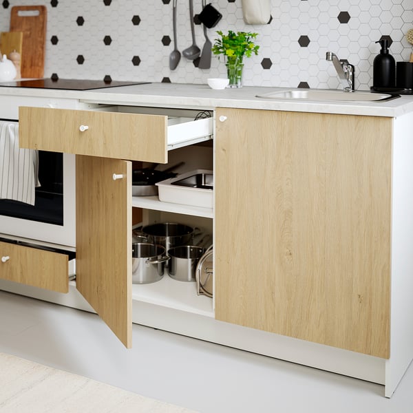 Modern kitchen with oak cabinets, open drawer, pots, oven, hexagonal tiles, sink, and hanging utensils.