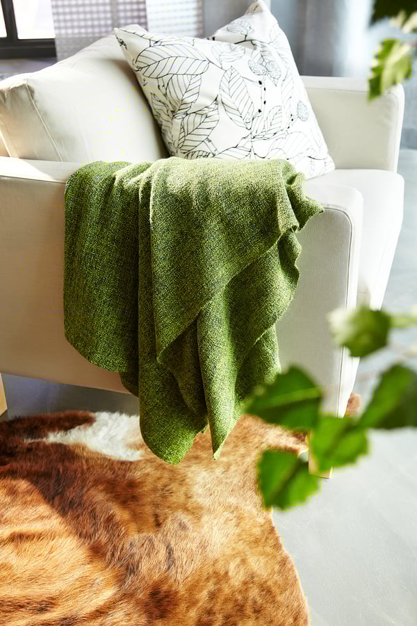 Cozy corner, beige couch with green throw blanket and leafy cushion, brown fur rug, plant nearby.