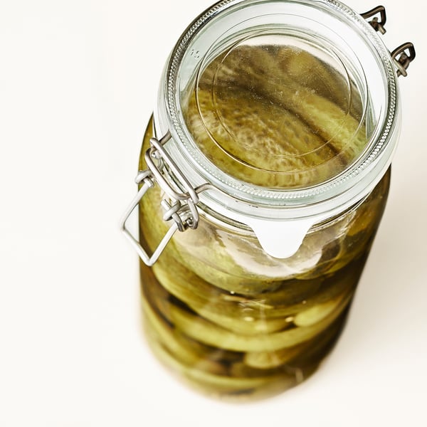 Open glass jar with lid and handle filled with pickles submerged in liquid, sealed airtight.