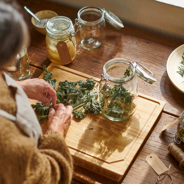 Person is pickling vegetables on a wooden chopping board. Glass jar labelled KORKEN with hermetic seal is used for preserving.