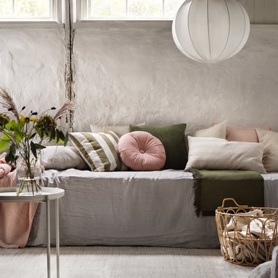 Cosy living room with grey KRANSBORRE round velvet cushion on white linen sofa, green throw, and woven basket on beige carpet.