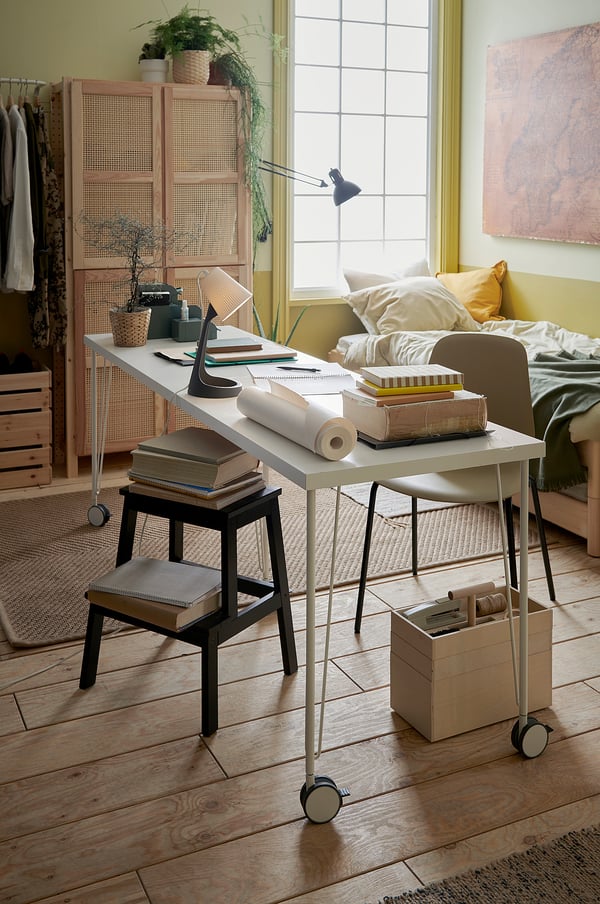 A bright room with a white KRILLE desk on wheels, a grey stool, and various items on top. Plants and clothes are nearby.