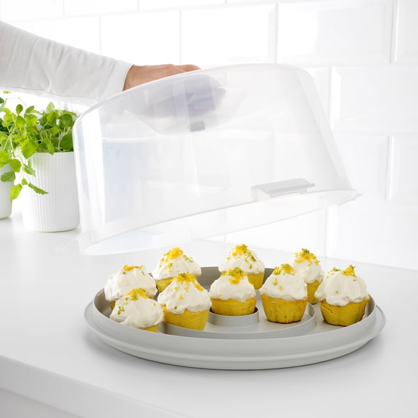 Hand holds clear lid of KRISPIG cake carrier, showing lemon cupcakes with icing on white countertop.