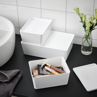 White KUGGIS boxes on bathroom counter, one stores makeup, another is empty, some stacked with lid on top. Grey towel and flowers in vase beside.