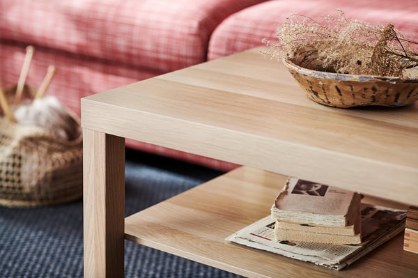A light wooden coffee table with a red couch and decorative basket behind.