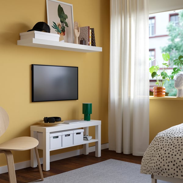Modern living room with yellow walls, white tv bench, flat-screen tv, green lamp, wooden chair, dotted armchair, large curtain, and plant.