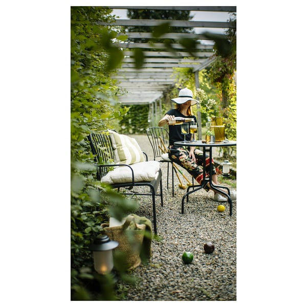 Person in garden diamonds patio, with LÄCKÖ black metal chairs, potted plants, table set with citrus fruits, glasses, and candles.