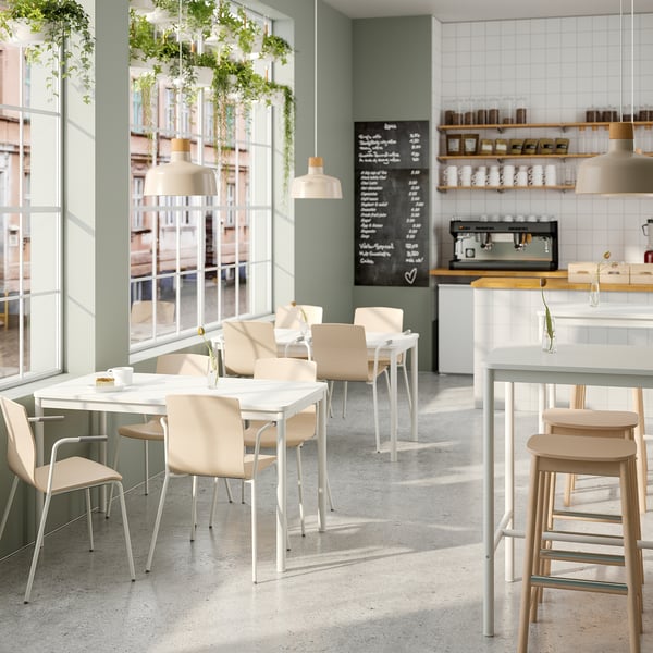 Modern café with white tables and light-colored chairs. Large windows, green walls, and hanging plants. Chalkboard menu and counter with coffees.