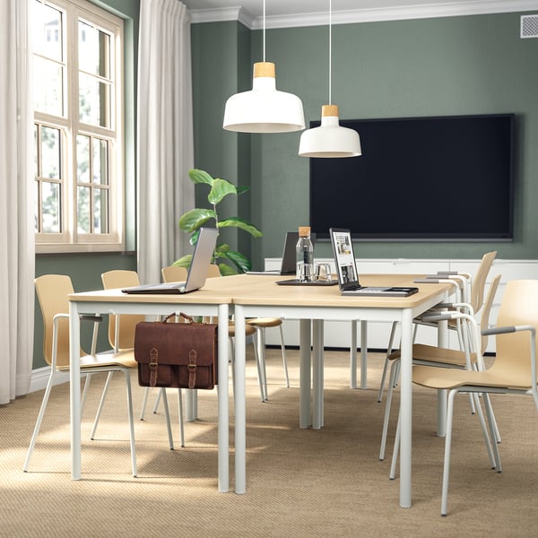 Modern office setup with wooden table, chairs, laptop, plant, 2 pendant lamps, tv, and window.