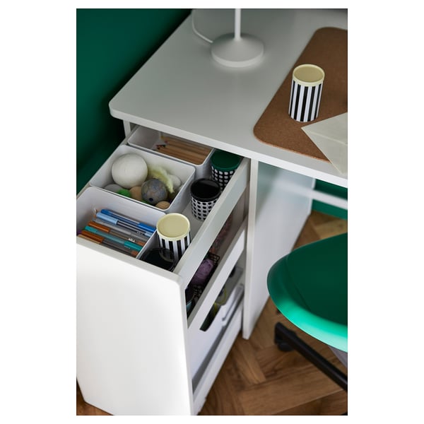 White desk with open drawer showing pens, books, and small toys. Green wall and matching chair nearby.