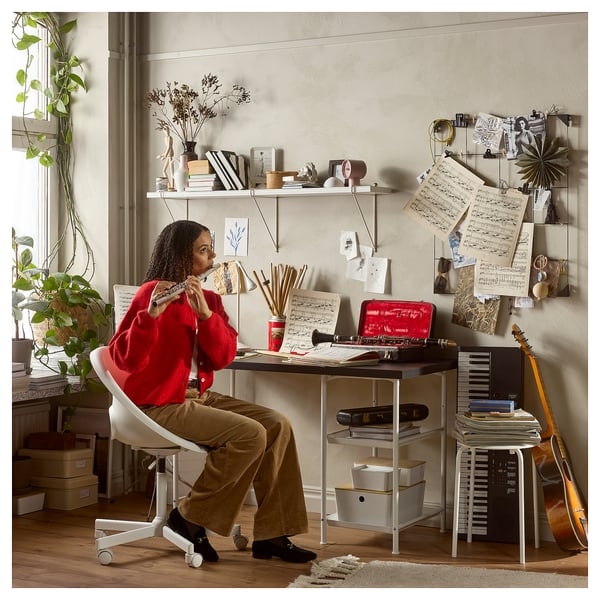 A person in a red sweater works at a white LAGKAPTEN desk with shelves, surrounded by musical instruments and decorated with notes and art supplies.