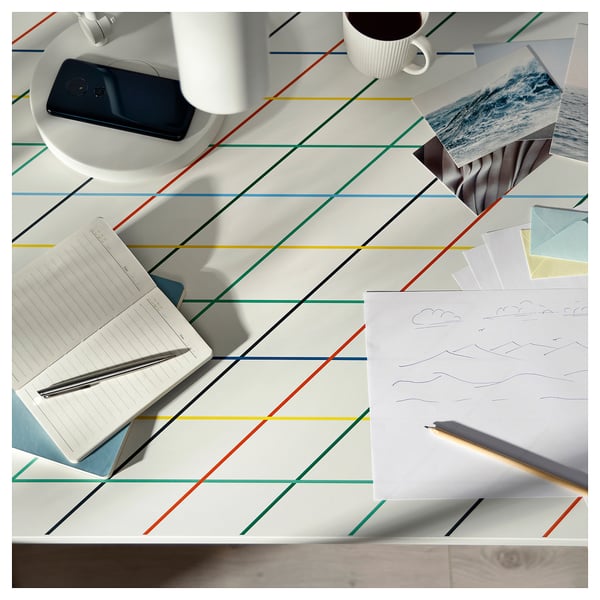 Scandinavian-style colourful grid-patterned white desk with notebook, phone, mug, and drawings.