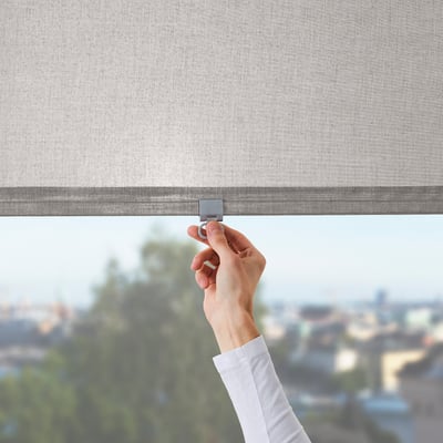A hand adjusts a LÅNGDANS roller blind, controlling light and privacy in a room with a city view.