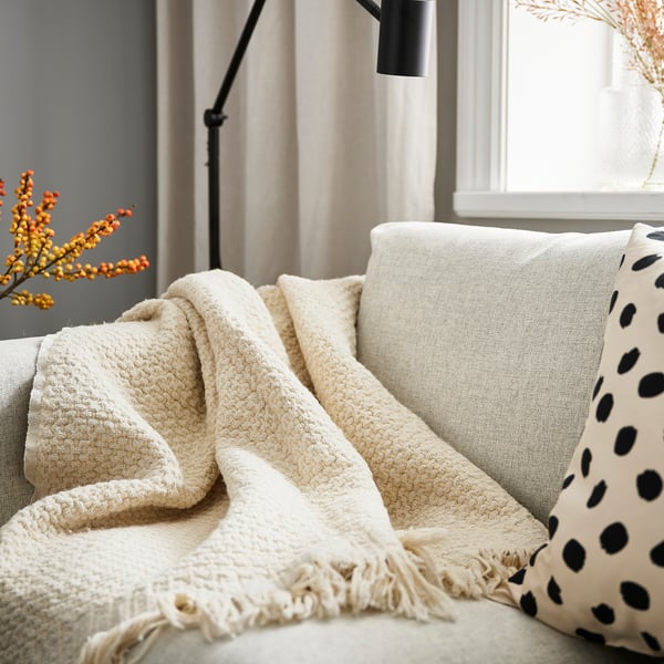 Off-white LAPPTÅG throw blanket, soft & warm, on sofa with black-spotted cushion.
