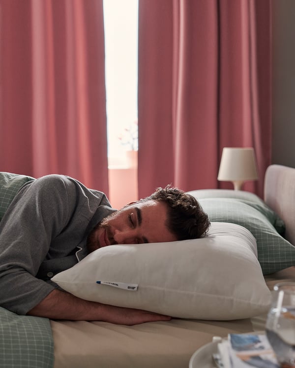 Person sleeps on LAPPTÅTEL grey pillow on bed with red curtains.