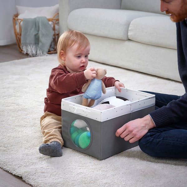 A child sits in a fabric storage box with a mesh window, holding a toy while an adult assists. The boxs handle aids in carrying or moving it.