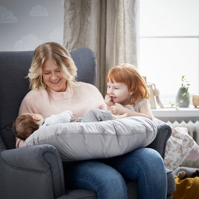 A person and two children enjoying a cosy moment on a grey couch with a nursing pillow, surrounded by soft decor in a well-lit room.