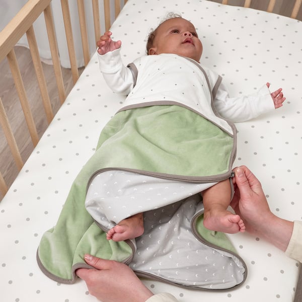 Baby in green sleep bag, zipped up, in cot being dressed.