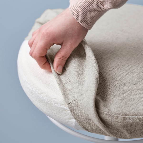 A hand adjusts a round, textured, beige LIDKULLEN sitting support on a silver base.