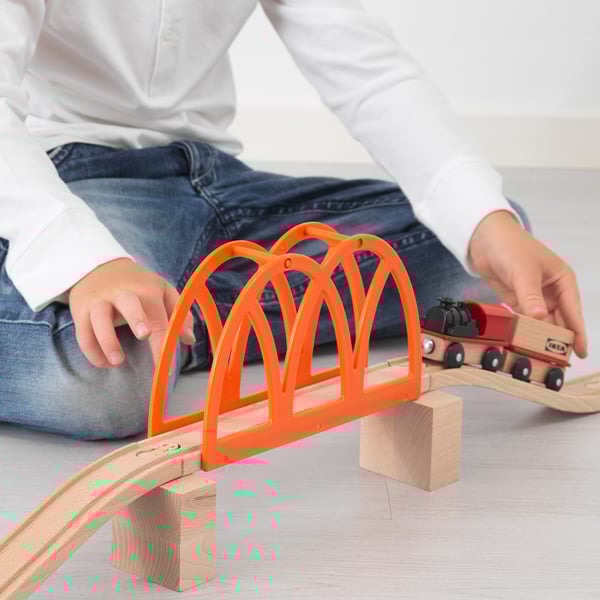 Child sitting on floor, playing with wooden train set. Bright orange bridge spans tracks, train crossing it, promoting imagination and motor skills.