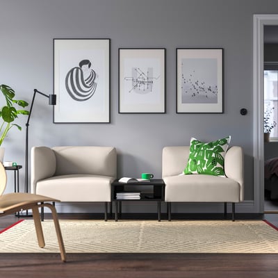 Modern living room with neutral sofas, green pillows, black coffee table, abstract art, grey walls, wooden floor, rug, and tall lamp with plant.