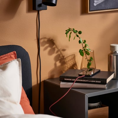 Bedside setup with a black LILLHULT usb cable charging a phone on top of books, near a plant and a wall lamp.