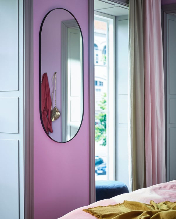 Modern bedroom with LINDBYN mirror reflecting red accessories on lavender wall, pink curtains, natural light.