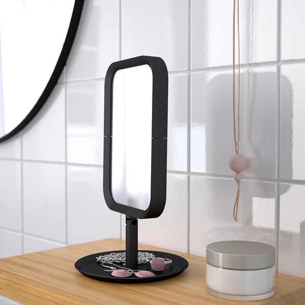 Addressable mirror with magnifying side, on wooden desk by tiled wall. Decorative items nearby.