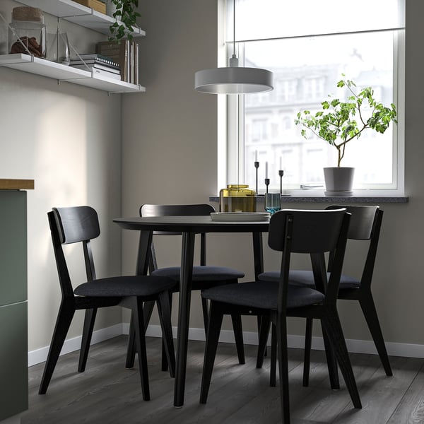 Four black chairs under a dark table, near a window with a plant and pendant light, modern dining setup.