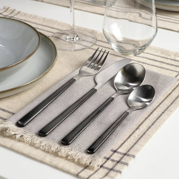 Modern table setting with LÖFTESRIK cutlery on beige placemat. Includes fork, knife, spoon, and empty wine glass.