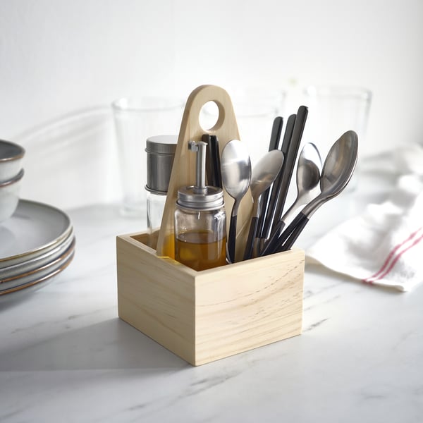 Wooden LÖFTESRIK cutlery caddy holds black-handled, stainless steel cutlery and small glass containers on a table with plates and glasses.