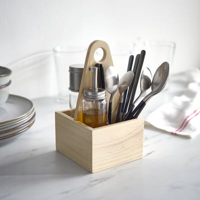 Wooden LÖFTESRIK cutlery caddy holds black-handled, stainless steel cutlery and small glass containers on a table with plates and glasses.