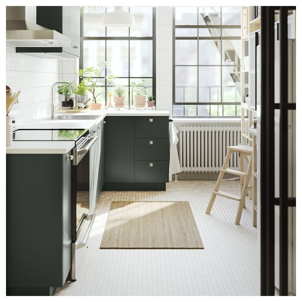 Modern kitchen with large windows, green cabinets, white countertops, wooden stools, and a beige jute rug on the tiled floor.