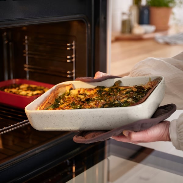 Person using oven gloves holds a large casserole dish with baked food, ready to place it in an oven, showcasing the dishs function for cooking meals.