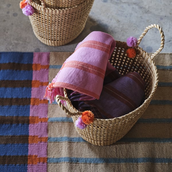 Two woven baskets with colourful pom-poms hold folded blankets on a striped rug.