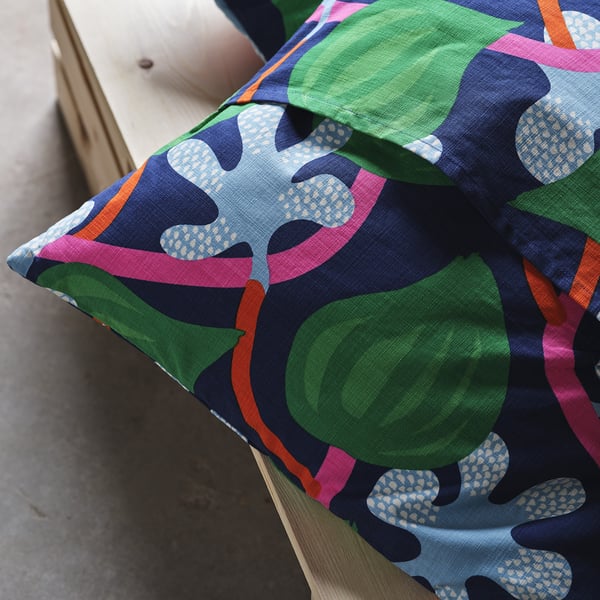 Brightly patterned cushion cover with green apples and pink leaves on a wooden chair. Front hand-embroidered caterpillar detail.