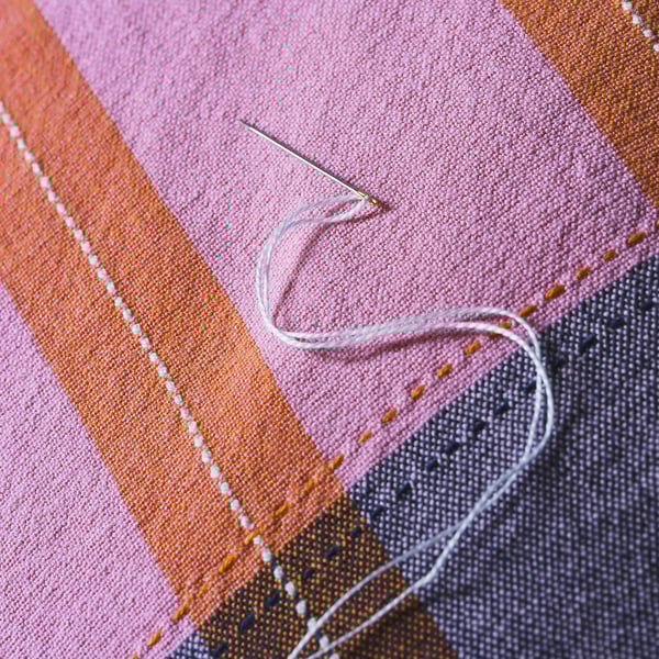 Close-up of handwoven fabric with needle and thread, showcasing intricate craftsmanship. Vibrant pink and orange stripes with detailed stitching.