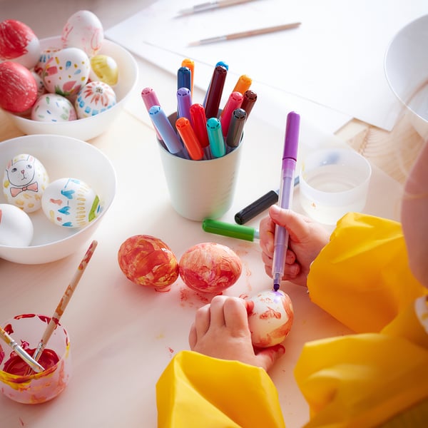 Creative scene with colourful easter eggs and drawing tools. Hands paint using vibrant pens. Yellow protective cover nearby.
